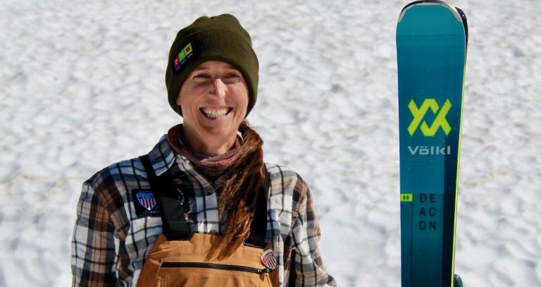 A woman smiles for the camera, standing next to a snowboard